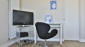 A modern room with a flat-screen TV on a stand, a black office chair, a glass desk, and a door.