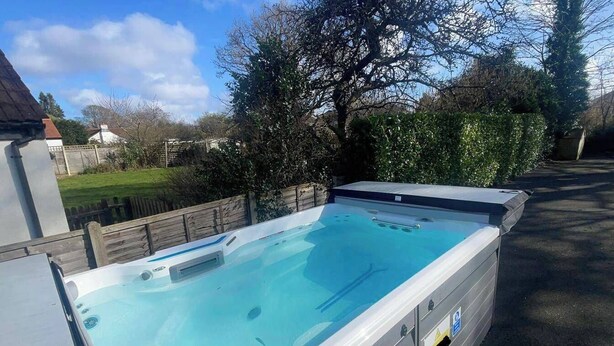 A hot tub with a clear blue interior, surrounded by a wooden deck and a hedge.