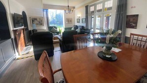 A dining area with a wooden table, chairs, and a vase of flowers. There are windows with curtains, a chandelier, and a television on a stand.