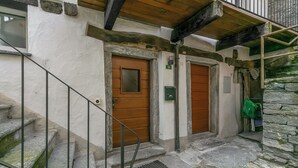 Un escalier en pierre mène à une porte en bois ornée d’une plaque numérotée, avec une main courante en métal et un petit sac vert accroché au mur.