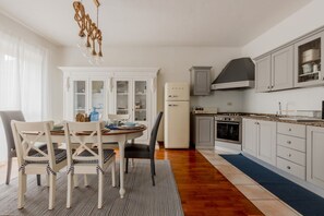 A kitchen with white cabinets, a white refrigerator, and a stove. There is a dining table with chairs and a chandelier.