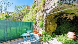 Eine Terrasse mit Tisch und Stühlen, ein grüner Zaun und eine Felswand mit einer höhlenartigen Öffnung.