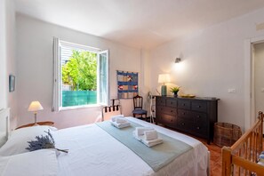 A bedroom with a bed, a dresser, a chair, a lamp, and a window with a view of greenery. - Yard Apartment with Balcony in Vernazza, Vernazza, Italy (Vernazza)