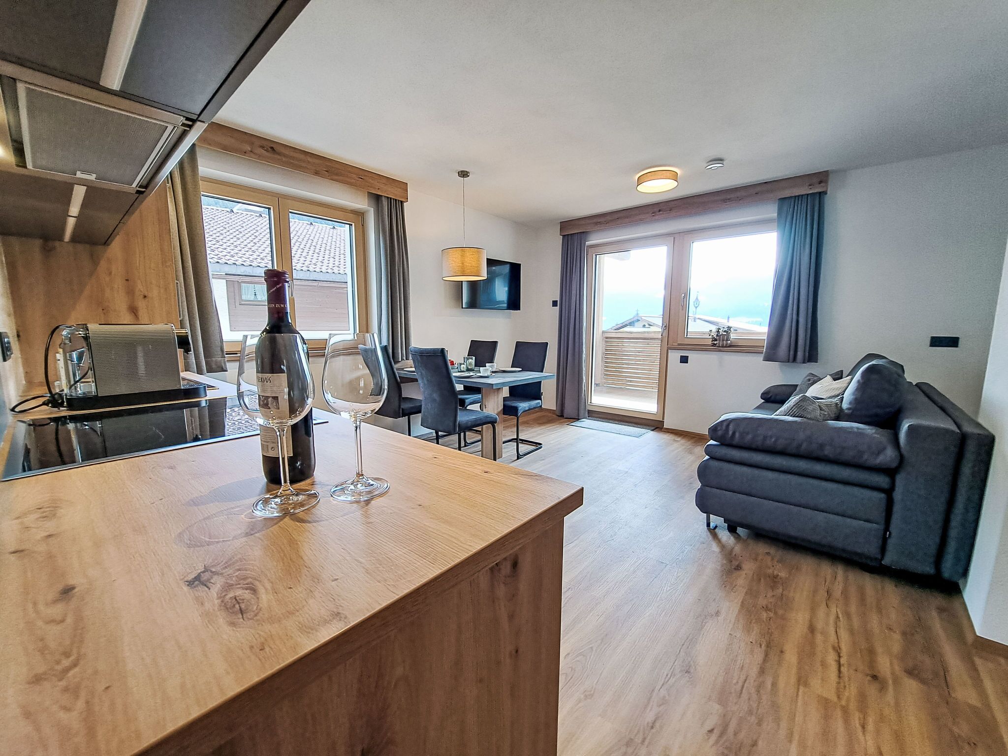 A modern living room with a wooden dining table, a sofa, and a kitchen area with a wine bottle and glasses.