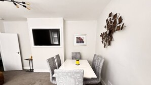 Un coin repas avec une table blanche et des chaises grises à motifs, une œuvre d’art accrochée au mur et une petite table d’appoint avec une plante.