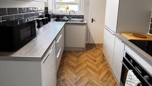 Une cuisine compacte avec un parquet en bois disposé en chevrons, un poêle noir et une fenêtre donnant sur les arbres.
