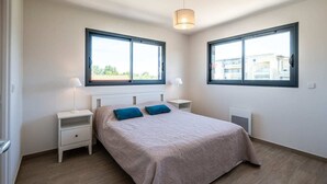 A bedroom with a bed, two bedside tables, a lamp, and two windows. - Maison Calypso de Narbonne (Narbonne)