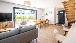A modern living room with a sofa, dining table, and chairs. There is a TV mounted on the wall and a staircase leading to another level. - Maison Calypso de Narbonne (Narbonne)