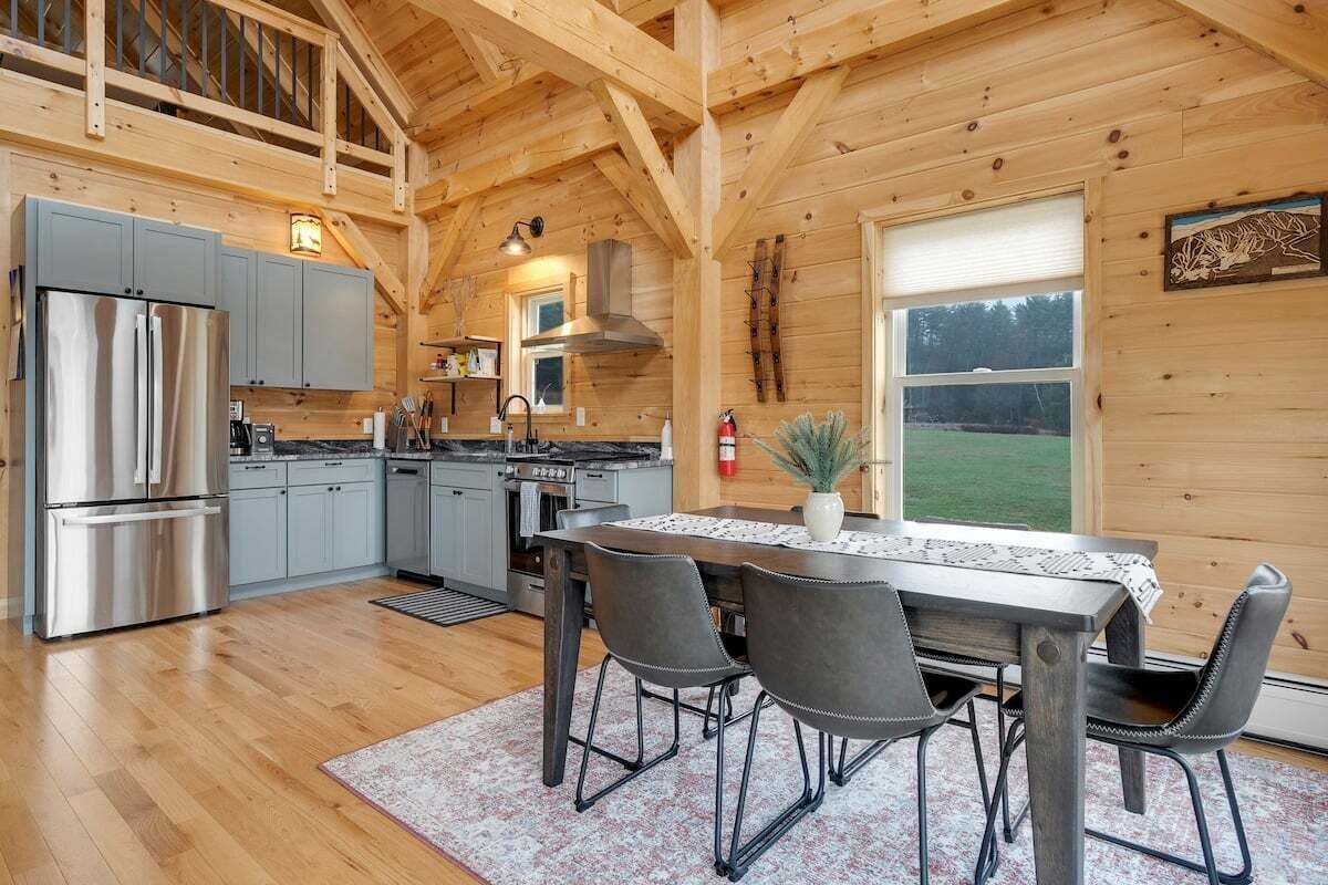 A wooden cabin interior with a kitchen, dining area, and large windows.