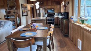 Un accogliente interno di una baita con un tavolo da pranzo in legno, sedie, una cucina con elettrodomestici moderni e un'ampia finestra con un modellino di una barca a vela.