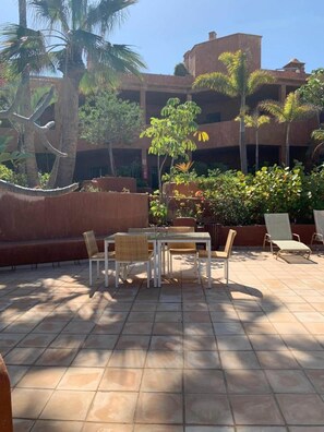 Un patio al aire libre con una mesa y sillas, rodeado de palmeras y un edificio con varios balcones.