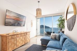 A modern living room with a large flat-screen TV, a blue sofa, a wicker console, and a balcony with a view of the sea.