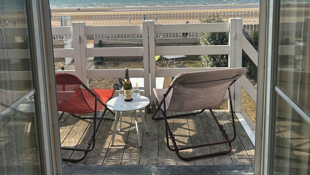 Un balcon avec deux chaises, une table avec une bouteille et des verres, et une vue sur la plage et l’océan.