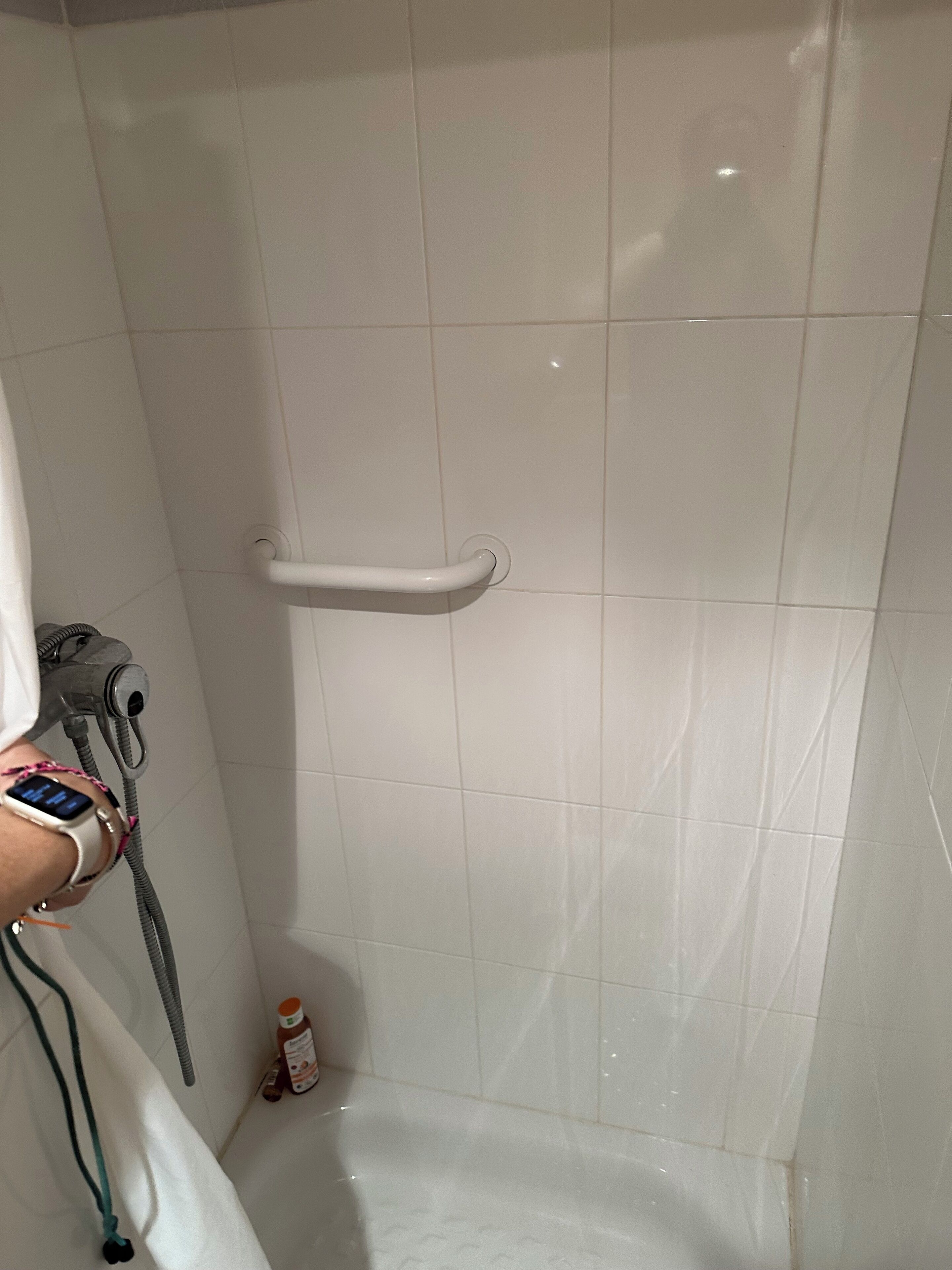 A bathroom with white tiles, a shower with a white handle, a hairdryer, and a bottle of lotion on the floor.