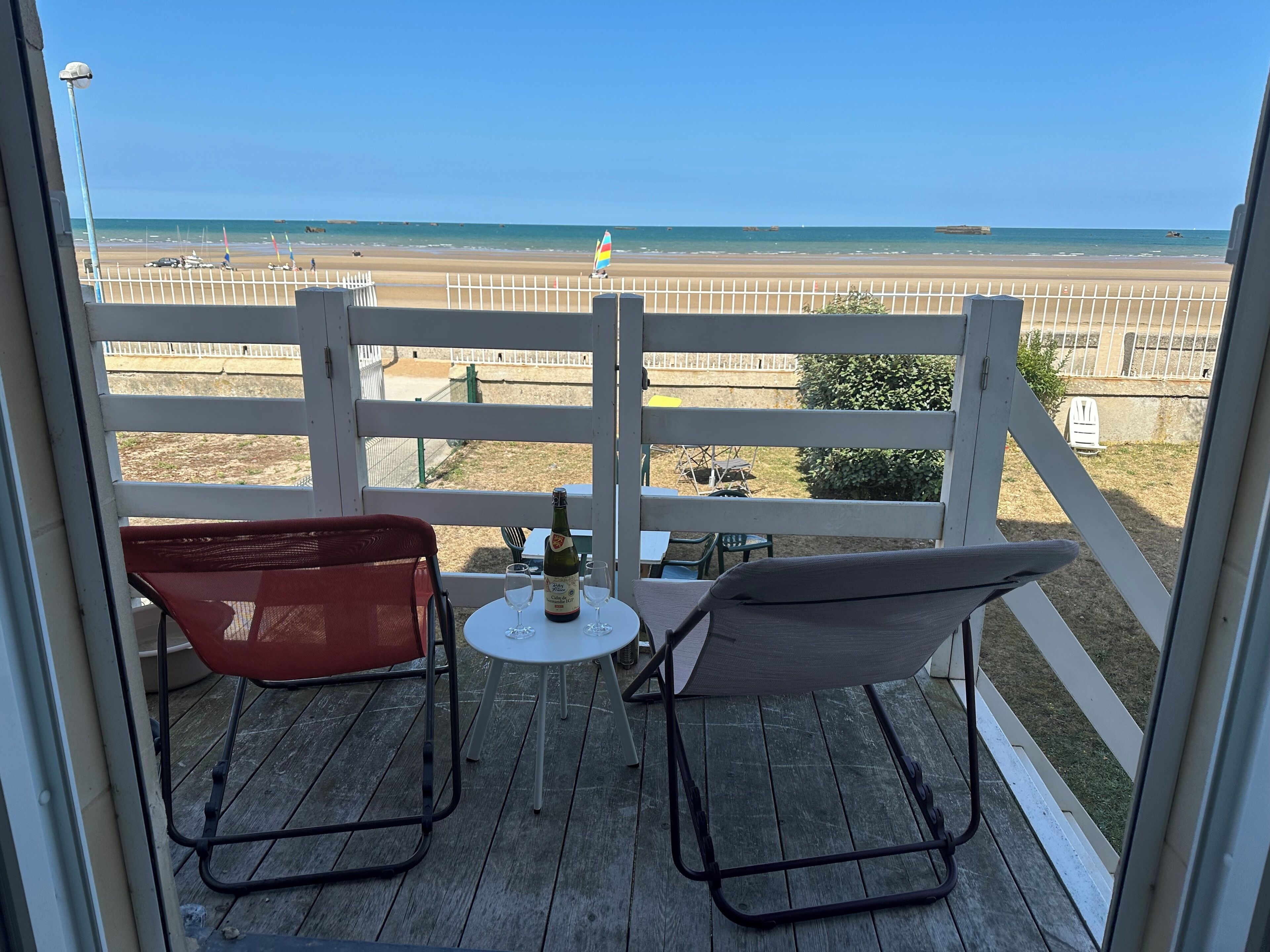A balcony with two chairs, a small table, and a bottle of champagne. The view includes a beach and a fence.