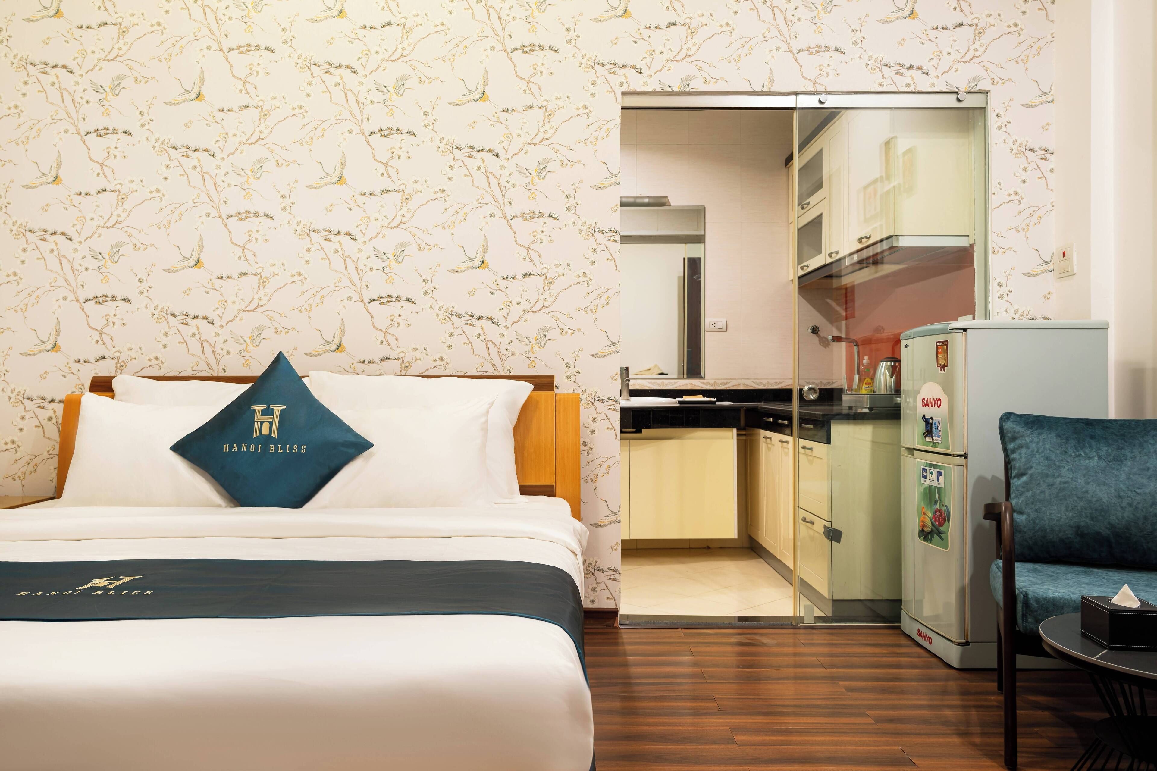 A hotel room with a bed, a blue pillow with a logo, a small kitchenette, and a blue chair.