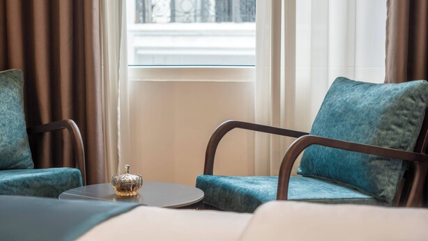 Ein Zimmer mit zwei blauen Polsterstühlen, einem kleinen runden Tisch und einem Fenster mit Vorhängen.