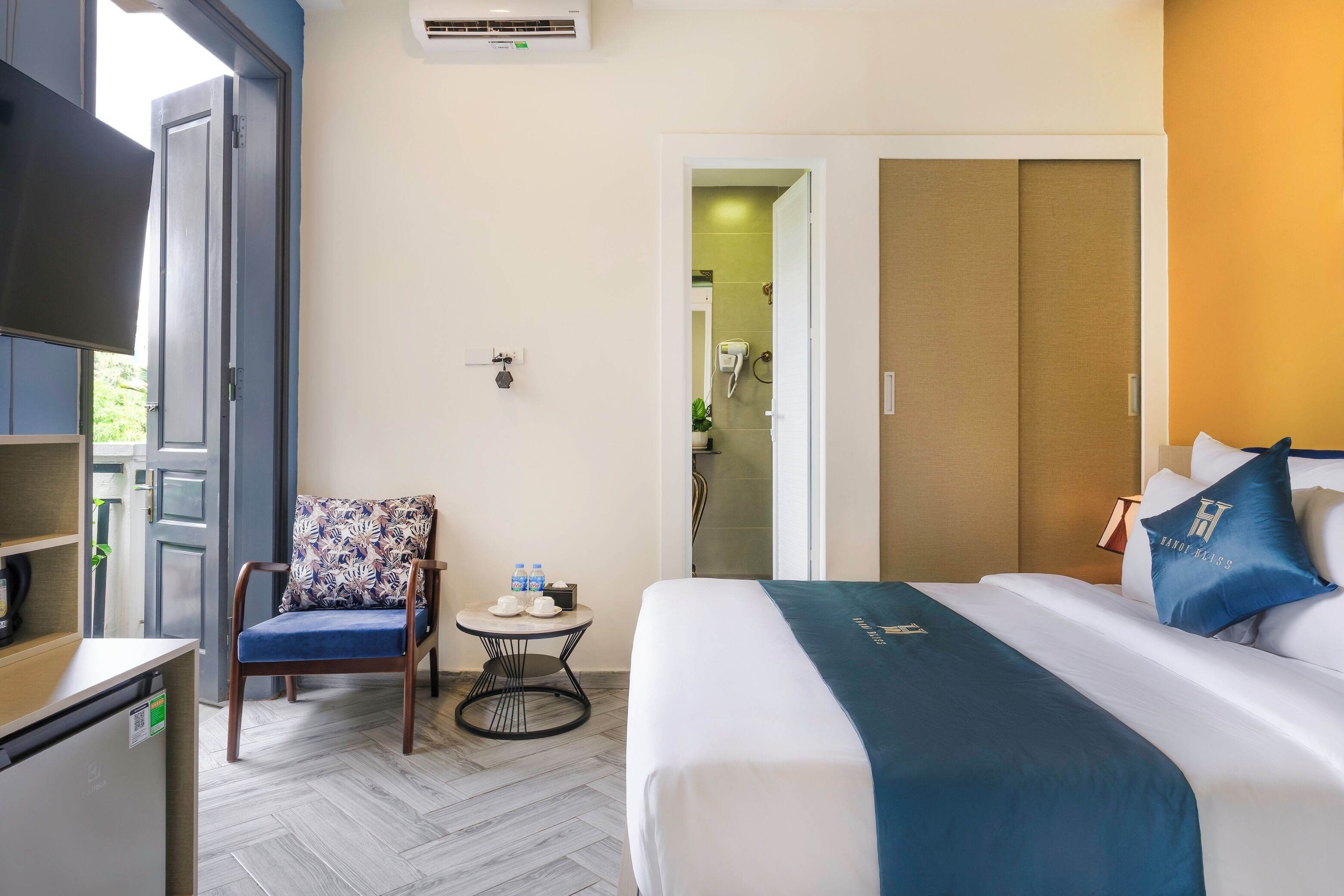 A hotel room with a bed, a chair, a small table with water bottles, a desk with a computer, and a wardrobe.