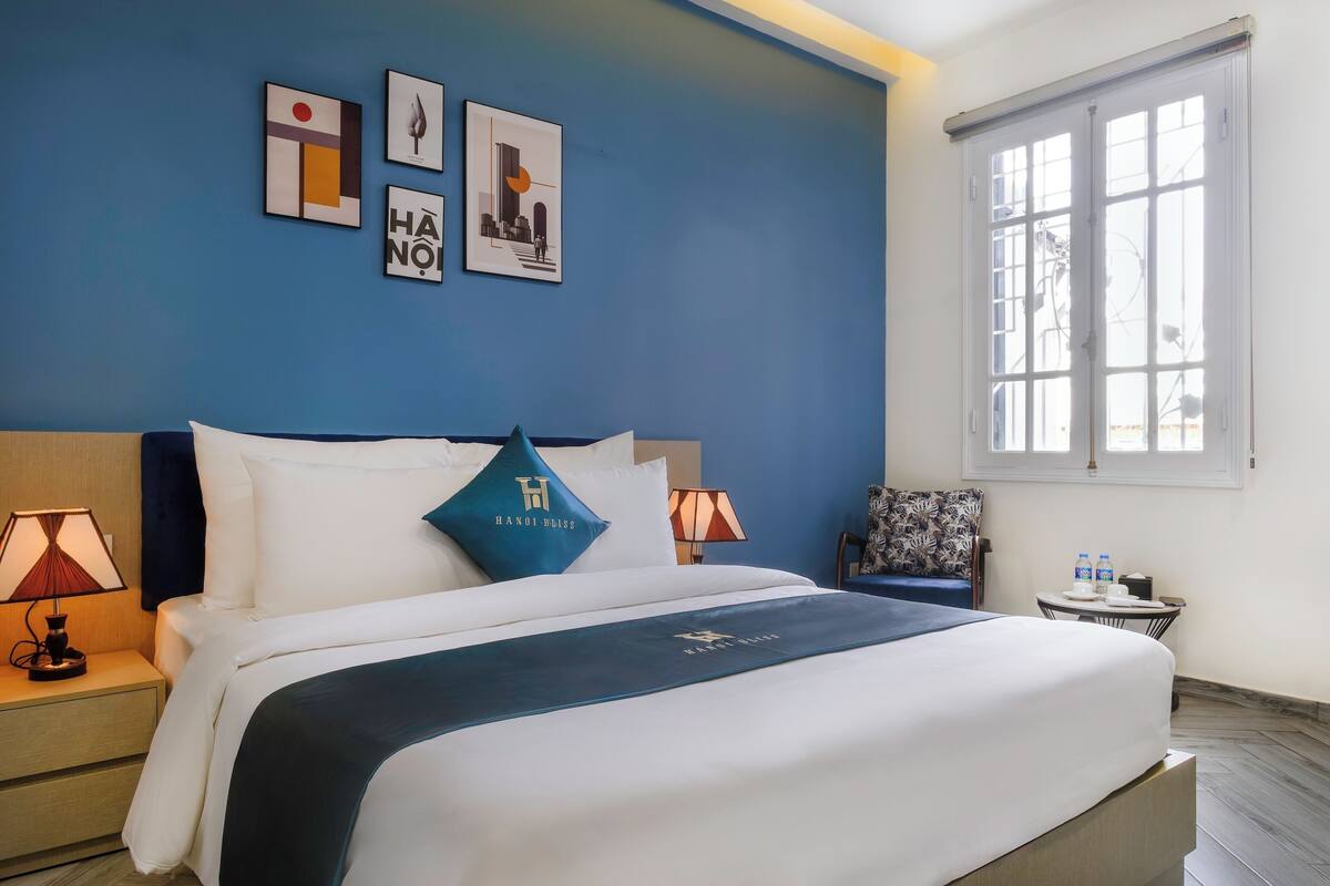 Une chambre d’hôtel avec un grand lit, une table de chevet avec une lampe, une petite table avec une chaise et une fenêtre donnant sur un beau paysage.