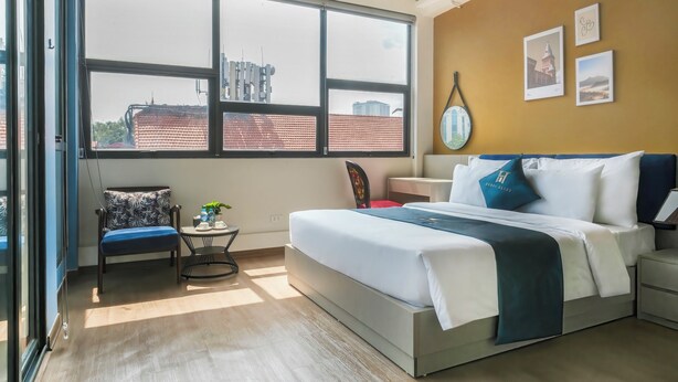 Een moderne slaapkamer met een groot raam, een bed met witte beddengoed en blauwe accenten, een klein rond tafeltje met een plant en een stoel met een bloemenprint.