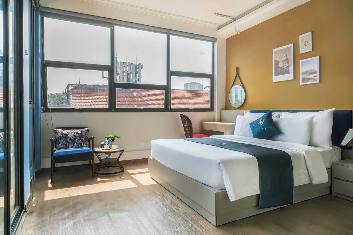 Une chambre moderne avec une grande fenêtre, un lit avec une literie blanche et des touches de bleu, une petite table ronde avec une plante, et une chaise à motifs floraux.