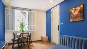 Een kamer met een eettafel en stoelen, een blauwe bank, een raam met een metalen hekwerk en een schilderij aan de muur.