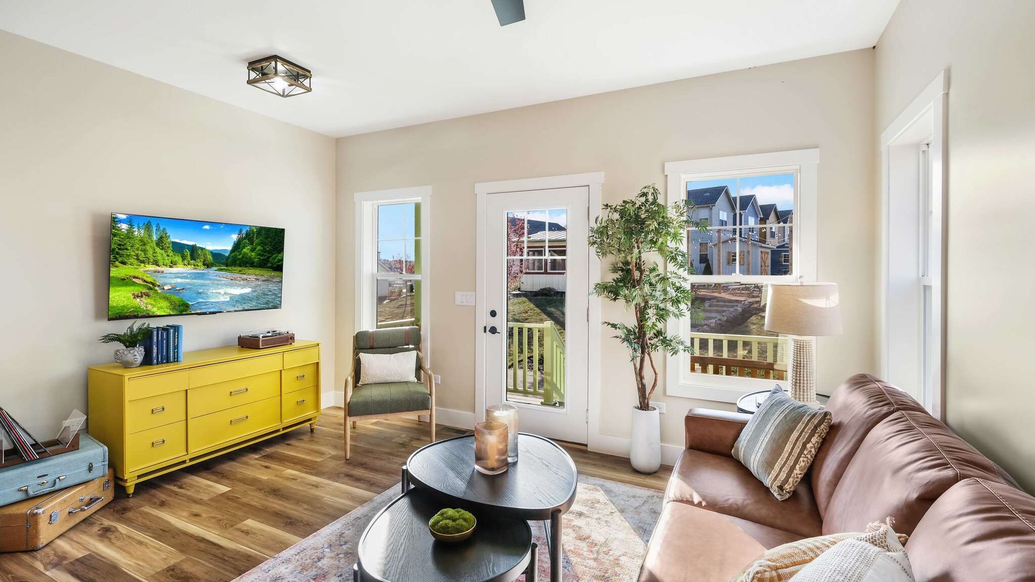 A living room with a TV, yellow dresser, leather couch, and a view of a house through a door.