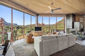 A spacious living room with a large gray sectional sofa, a flat-screen TV, and a fireplace. The room features large windows offering a scenic view of red rock formations and greenery.
