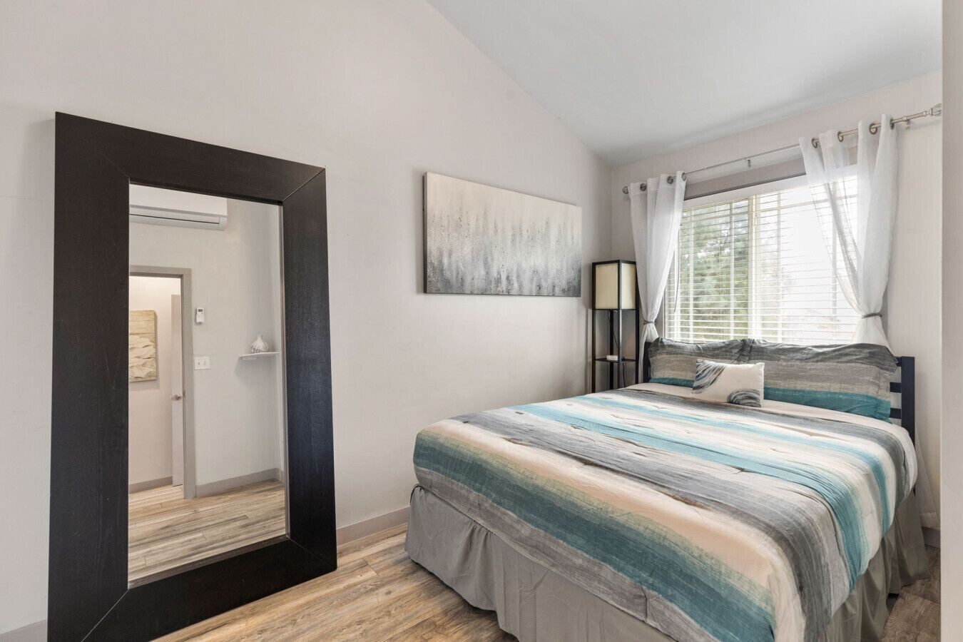 A bedroom with a bed, a large mirror, a painting, a bedside lamp, and a window with curtains.