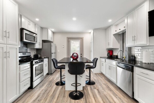 A modern kitchen with white cabinets, a central island with bar stools, stainless steel appliances, and a view through a glass door.