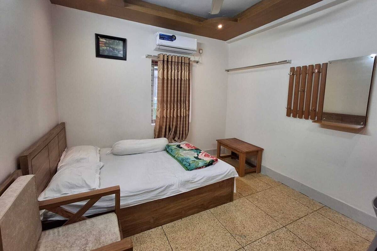 A bedroom with a wooden bed, a ceiling fan, an air conditioning unit, a small bedside table, a mirror, and a window with curtains.