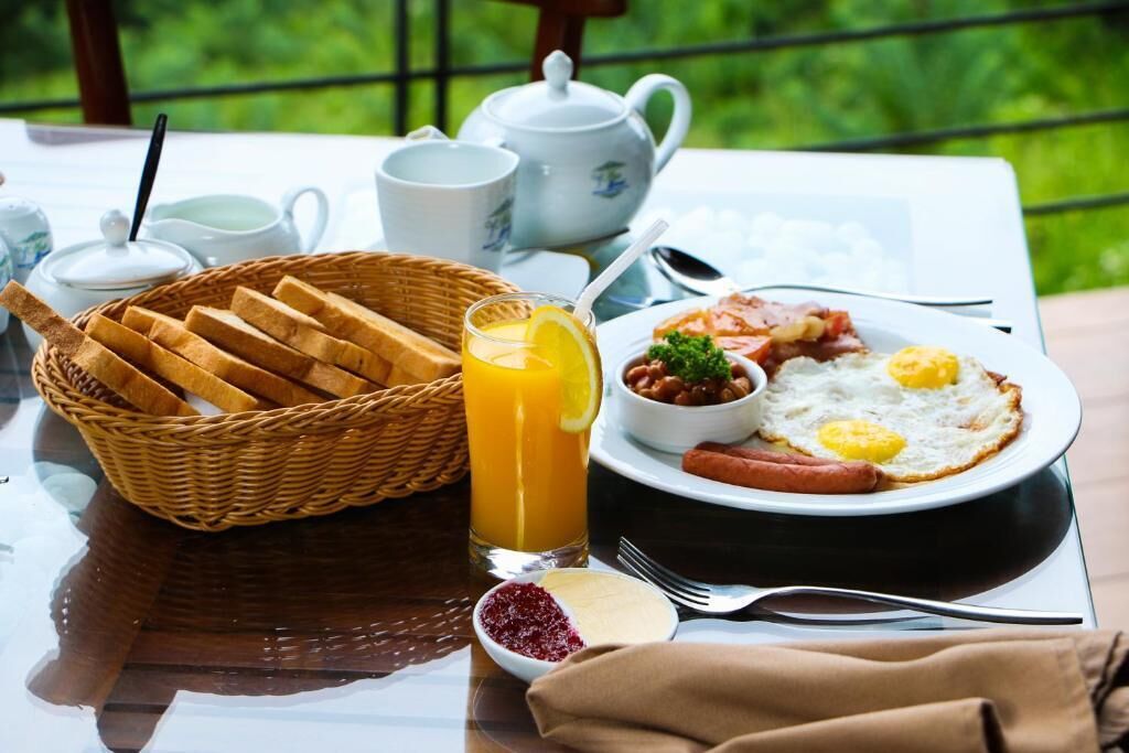 Ein Frühstückstisch mit Toast, Eiern, Würstchen und einem Glas Orangensaft, mit Blick auf eine grüne Landschaft.