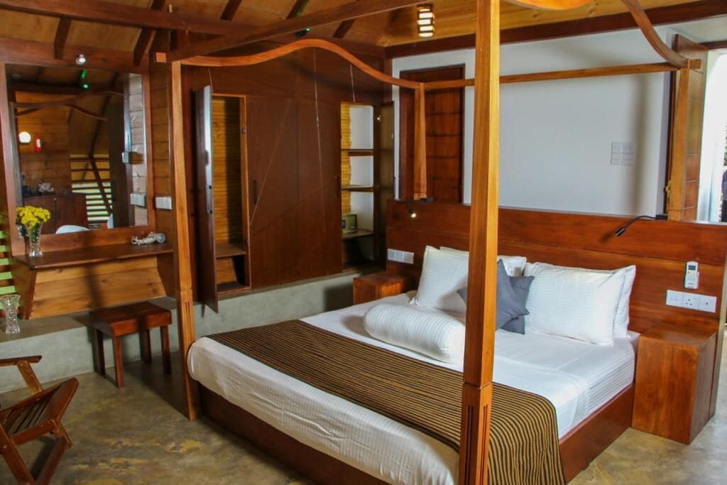 A wooden four-poster bed with white linens, a small wooden table with a vase of flowers, and a wooden bench.