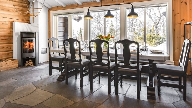 A cozy cabin interior with a stone fireplace, wooden dining table, and chairs, surrounded by large windows offering a view of trees. - Fjälldrömmen by Interhome (Storlien)