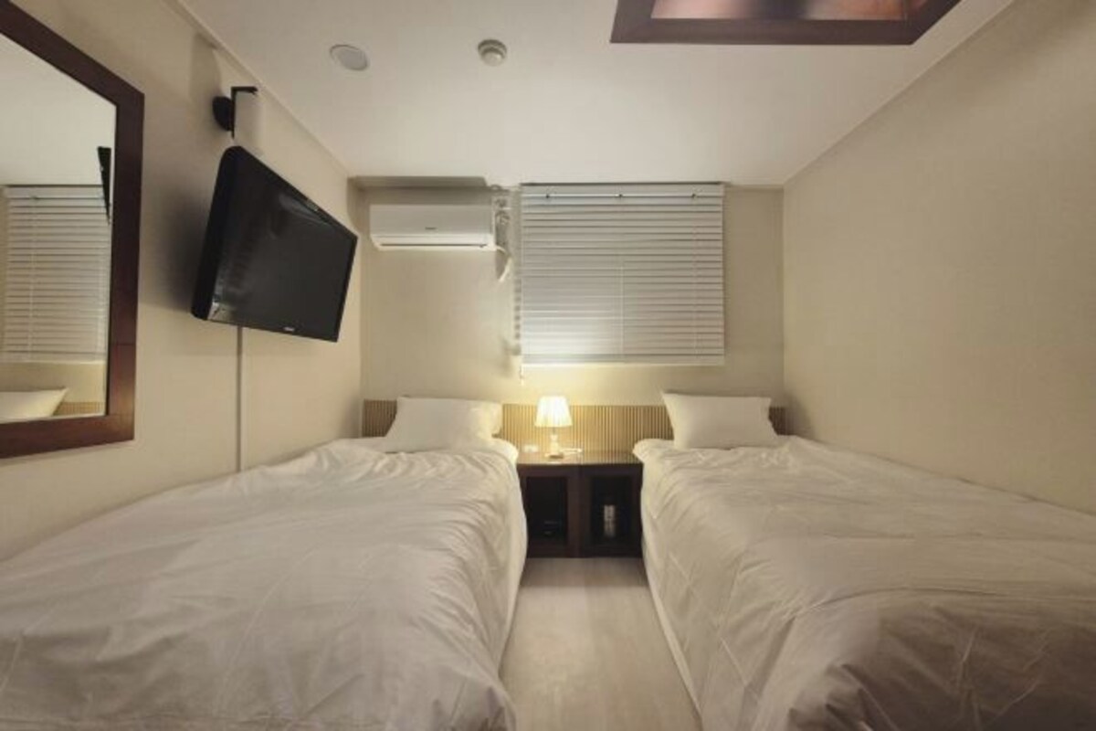 A hotel room with two single beds, a TV mounted on the wall, a window with blinds, and a nightstand with a lamp.