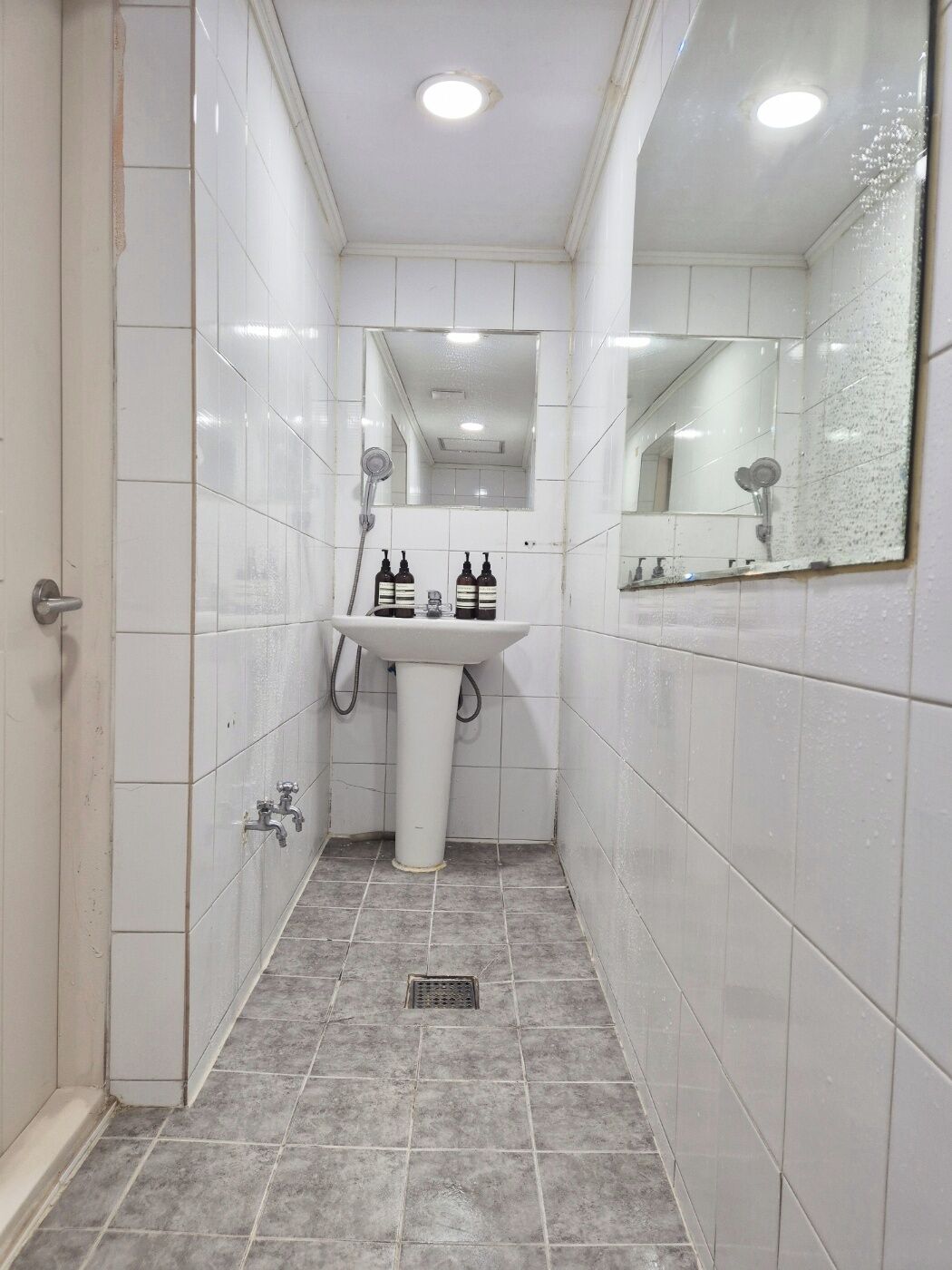 A narrow bathroom with white tiles, a white sink, two bottles of hand soap, a handheld showerhead, and a mirror.