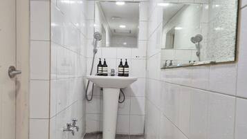 A narrow bathroom with white tiles, a white sink, two bottles of hand soap, a handheld showerhead, and a mirror.