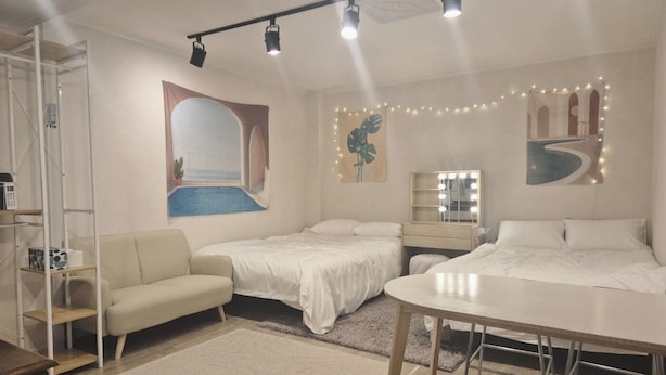 Une chambre avec deux lits, un bureau, une chaise, une coiffeuse avec éclairage, une grande fenêtre et une plante.