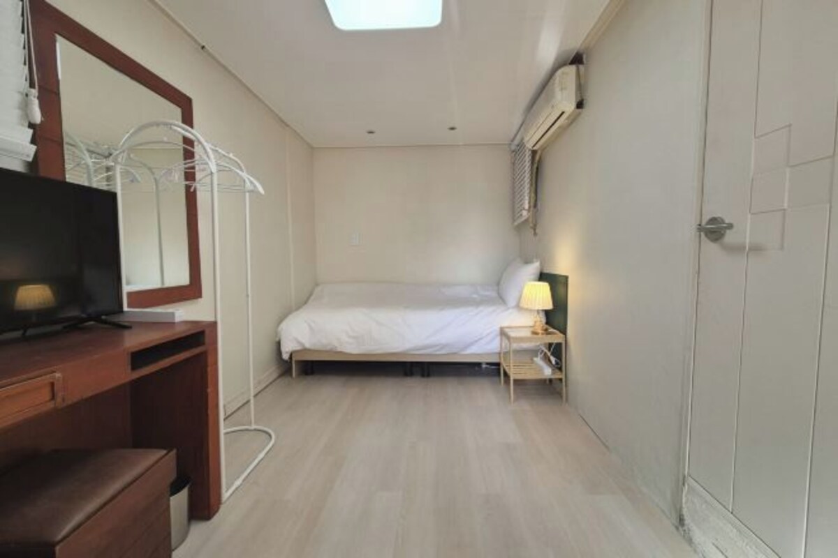 A small, well-lit bedroom with a bed, bedside table, lamp, and a TV on a wooden dresser.