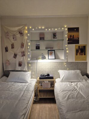 A bedroom with two beds, a bedside table with a lamp, a shelf with books and photos, and string lights.