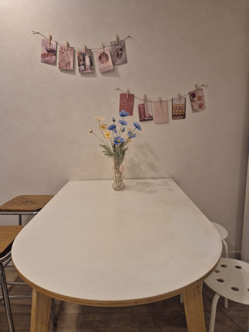Una mesa redonda con un florero, sillas de madera y una pared con pinzas sosteniendo fotografías.