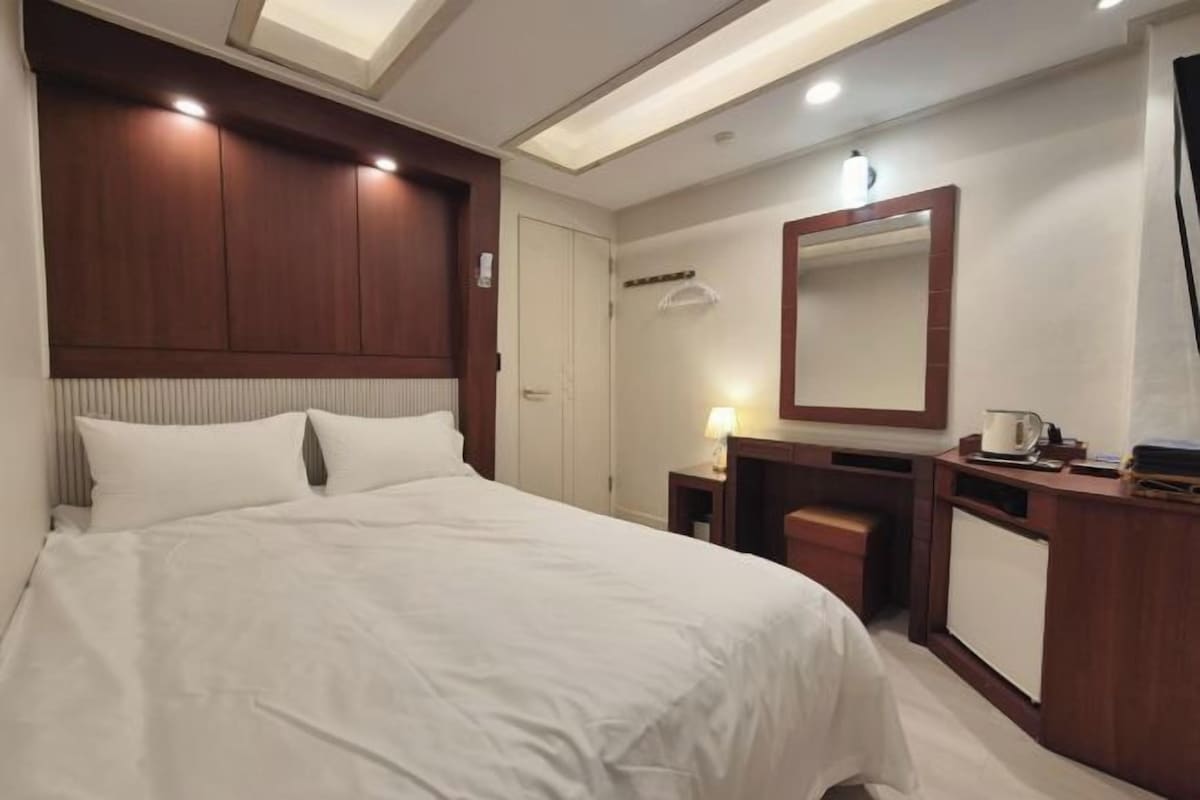 Una habitación de hotel con una cama grande, un cabecero de madera, una mesita de noche con lámpara, un espejo y un armario pequeño.