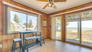 Quarto com decoração em madeira, ventilador de teto, mesa com dois banquinhos, janelas amplas com vista para a praia e porta de vidro deslizante para área externa.