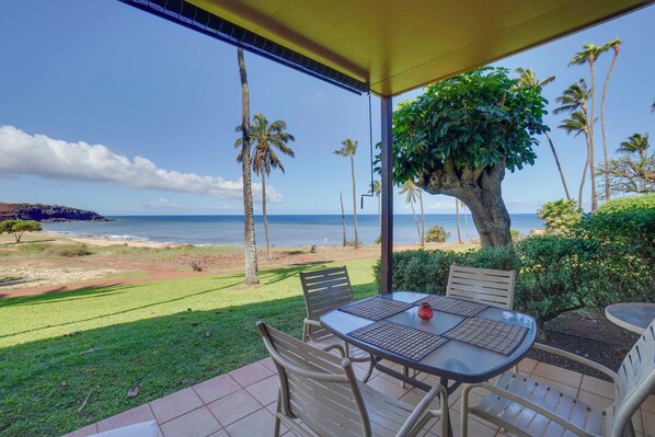 Un espace extérieur aménagé, abrité et doté de tables et de chaises, avec vue sur une plage bordée de palmiers et sur l’océan.