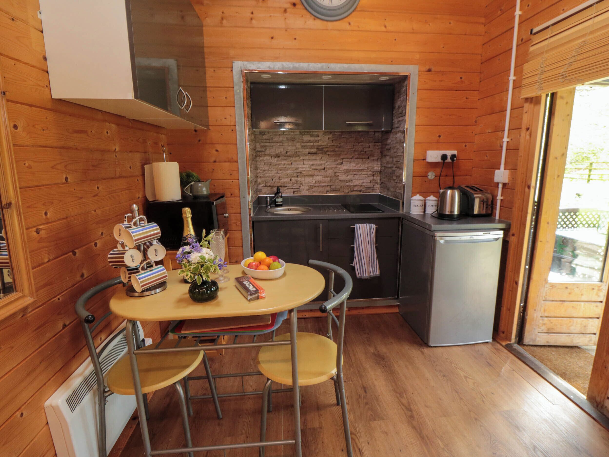 A compact kitchen with wooden walls, a built-in stove, a sink, and a small refrigerator. There is a dining table with chairs and a vase of flowers.