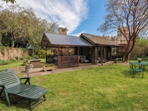 A covered outdoor seating area with a table and chairs, a barbecue grill, and a stone wall in the background. - Snape Castle Garden Lodge (Bedale)