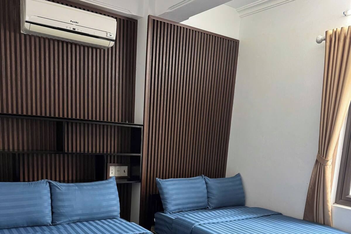 A hotel room with two beds, a built-in air conditioning unit, a wooden panel wall, and a window with curtains.