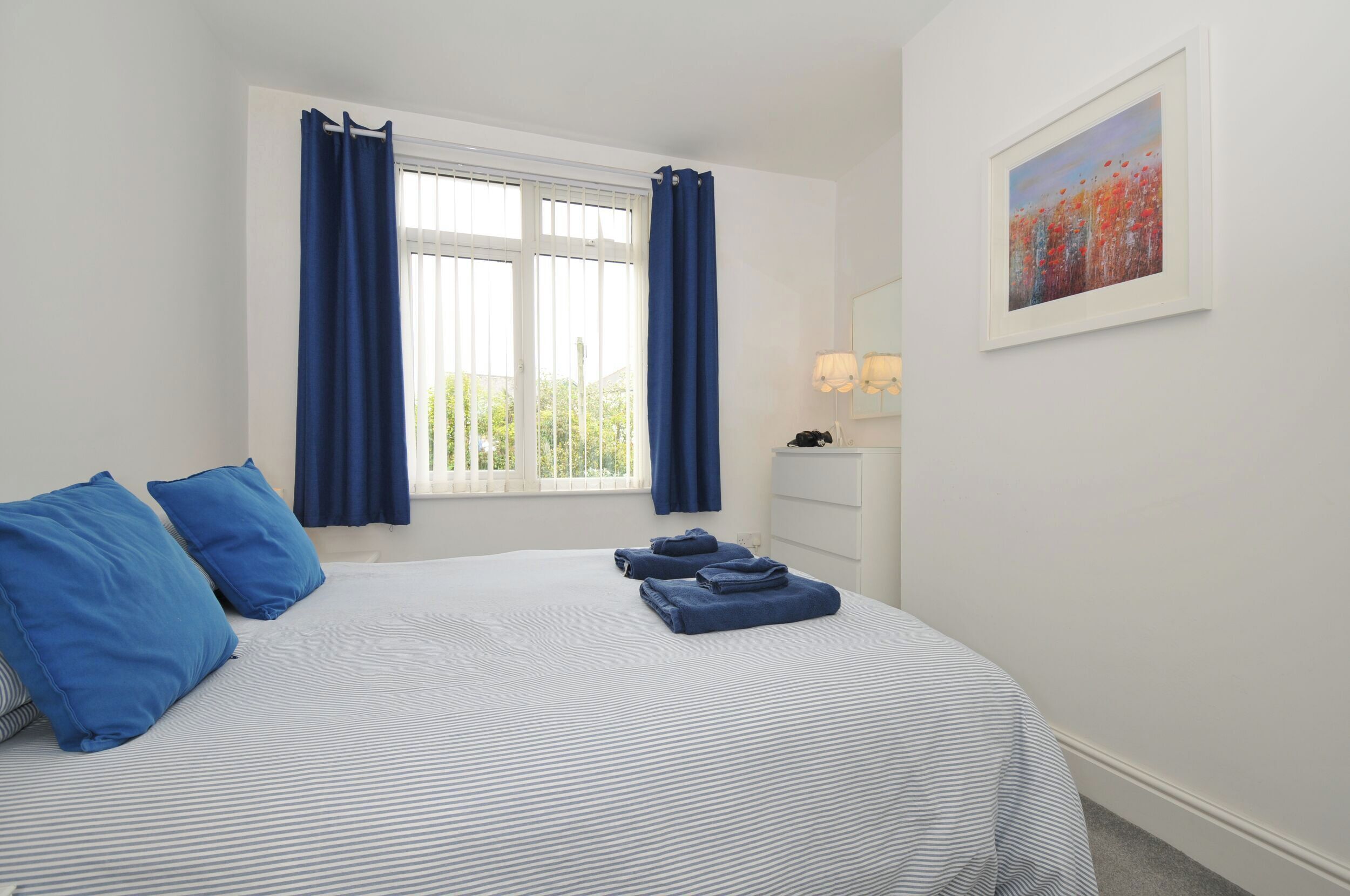 A neatly made bed with blue bedding, a window with curtains, a bedside table with a lamp, and a painting on the wall.