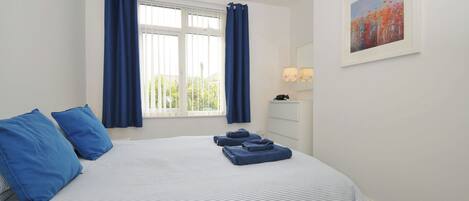 A neatly made bed with blue bedding, a window with curtains, a bedside table with a lamp, and a painting on the wall.