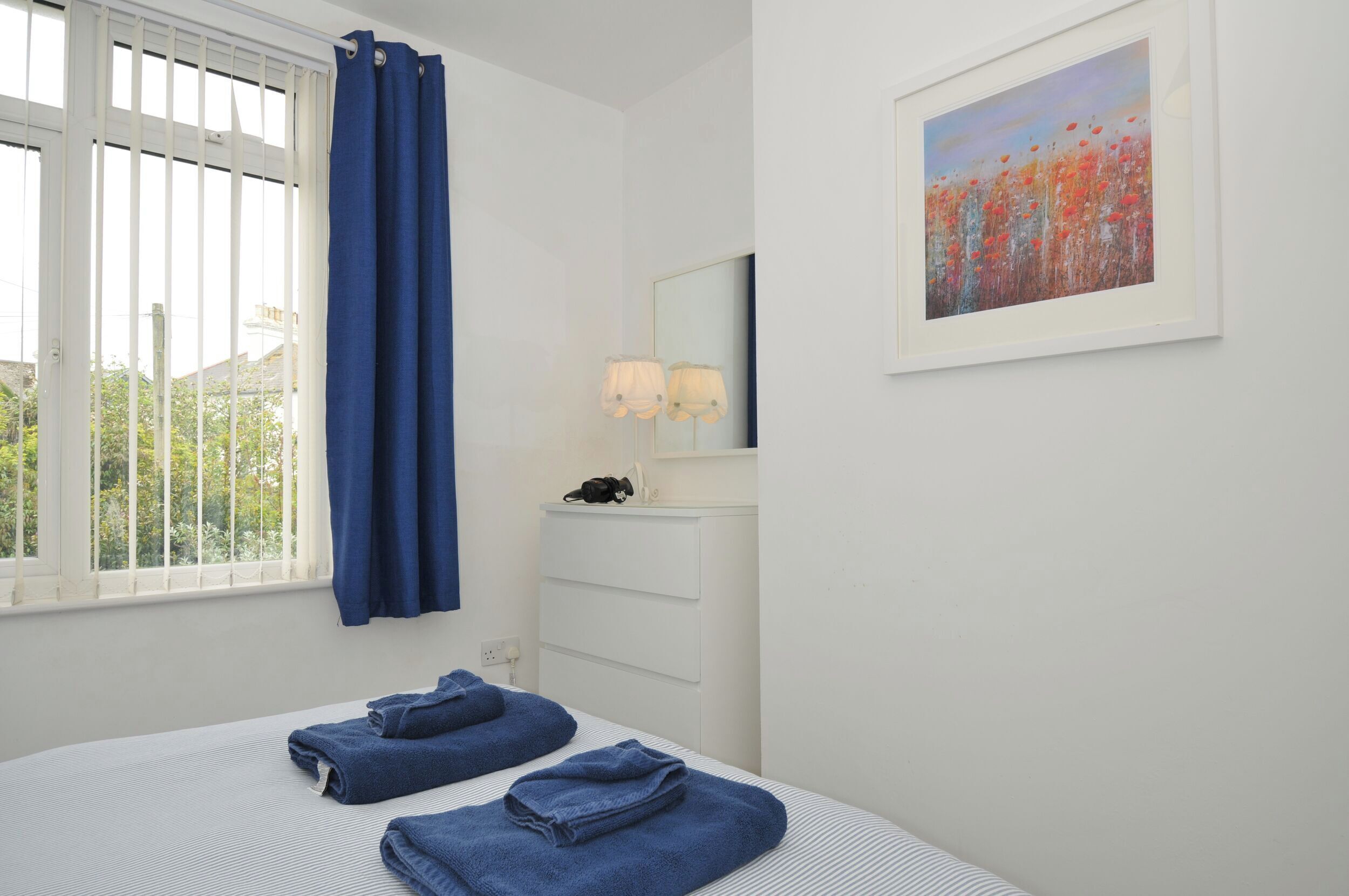 A bedroom with a bed, a nightstand, a lamp, a painting, and a window with curtains.